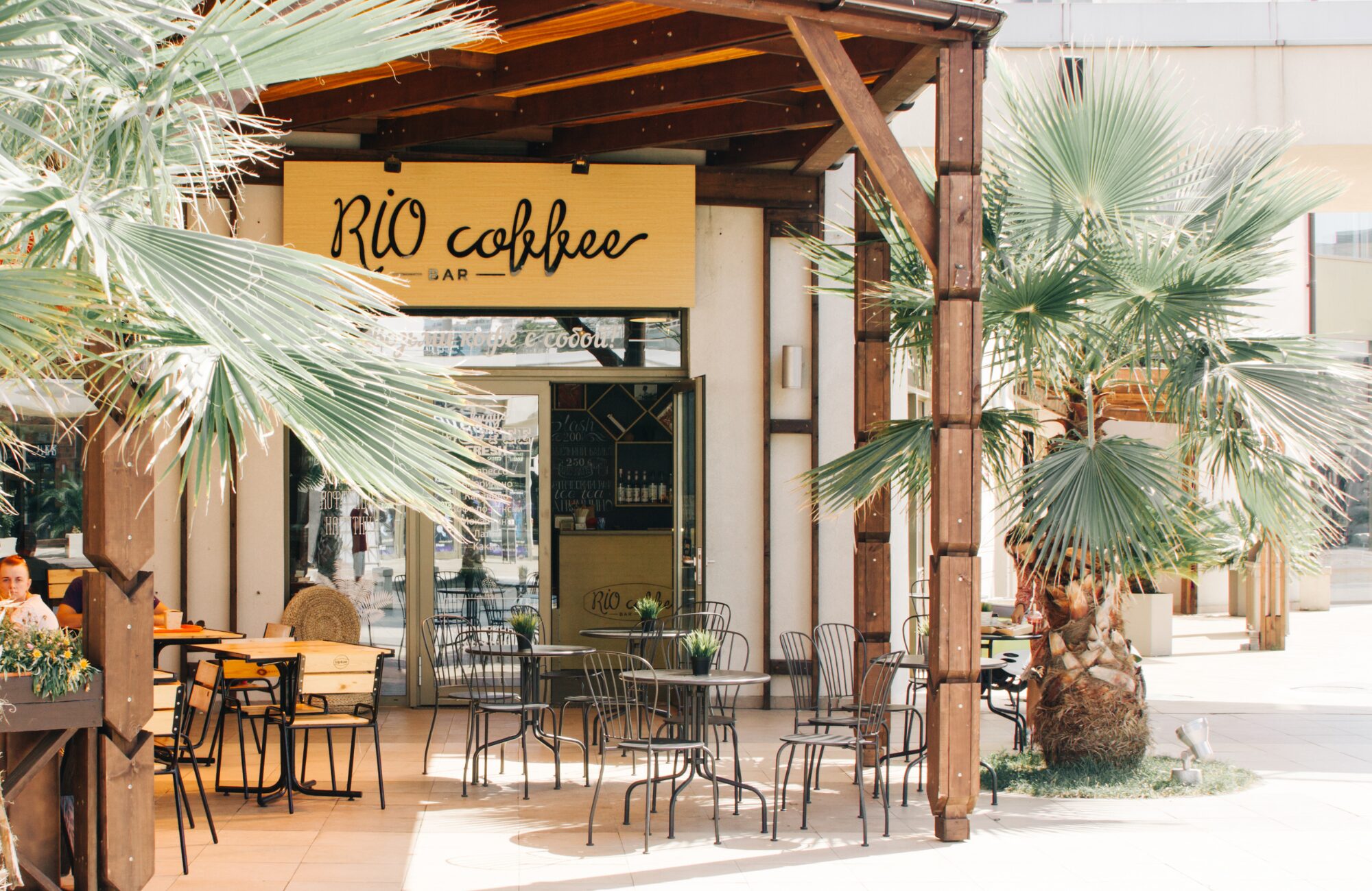 Outdoor café with a wooden canopy, metal chairs, and tables surrounded by palm trees, featuring a sign that reads "Rio Coffee.